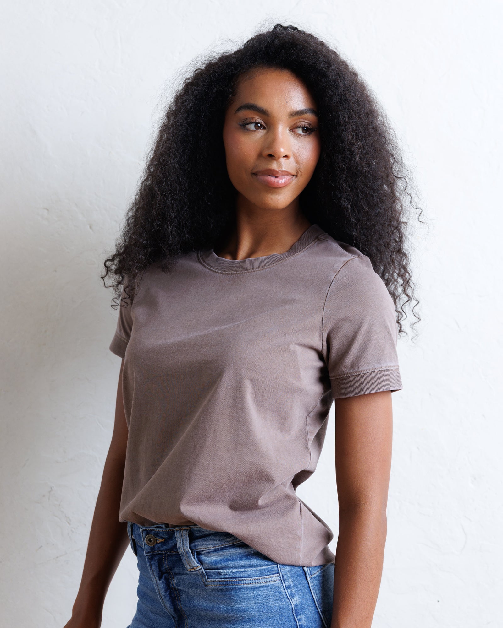 A woman with long, curly black hair wearing a muted taupe-brown short-sleeve crewneck t-shirt and blue jeans, standing against a white textured wall.