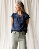 Woman wearing a blue denim top and light gray pants against a white curtain background