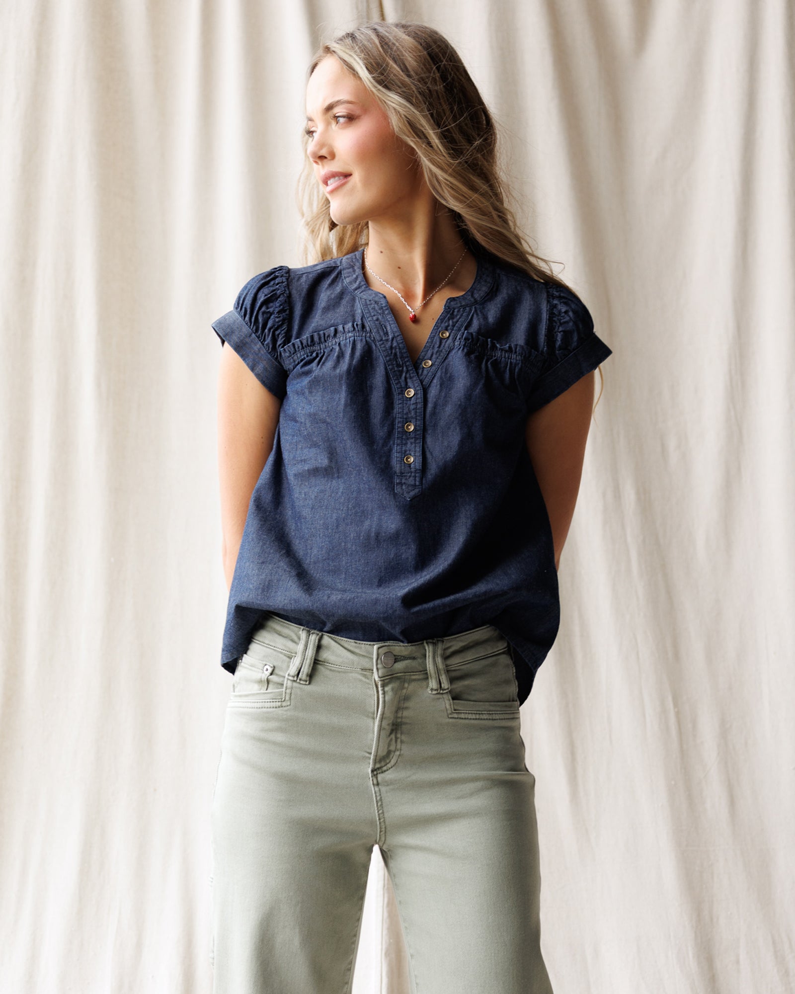 Woman wearing a blue denim top and light gray pants against a white curtain background