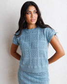 A woman wearing a light blue short-sleeve knit sweater with an openwork textured pattern, standing against a textured off-white wall.