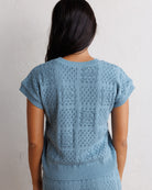 A woman viewed from behind wearing a light blue open-knit short-sleeve top with a textured geometric pattern, set against a plain off-white wall.