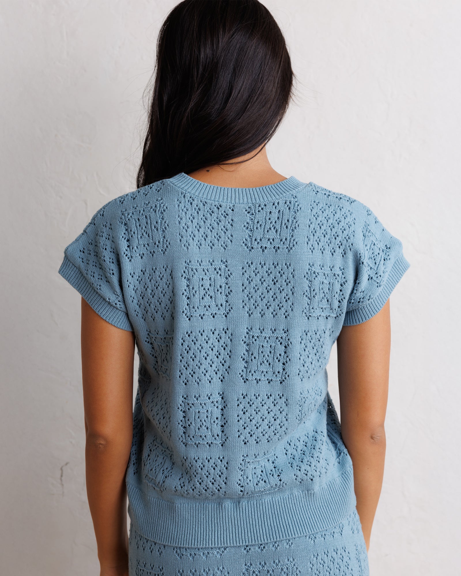 A woman viewed from behind wearing a light blue open-knit short-sleeve top with a textured geometric pattern, set against a plain off-white wall.