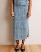 A person wearing a light blue open-knit midi skirt with a side slit, paired with black lace-up flats, standing against a textured off-white wall on a light wood floor.