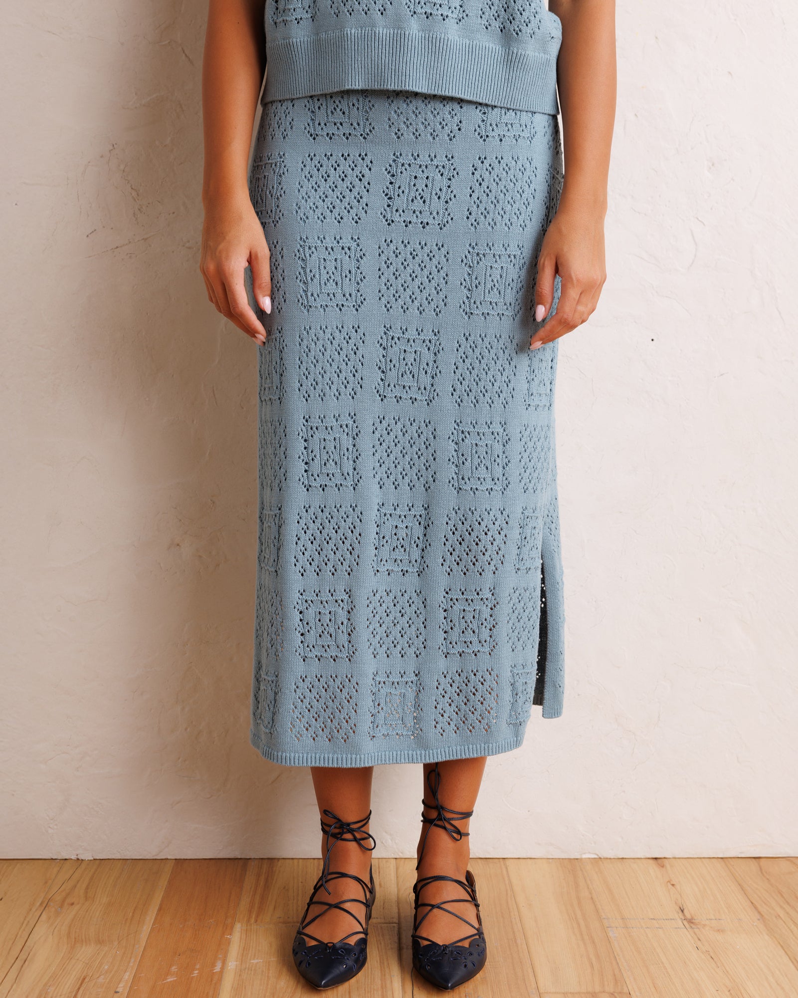 A person wearing a light blue open-knit midi skirt with a side slit, paired with black lace-up flats, standing against a textured off-white wall on a light wood floor.