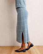 A person wearing a light blue open-knit midi skirt with a side slit, paired with black lace-up flats, standing against a textured off-white wall on a light wood floor.