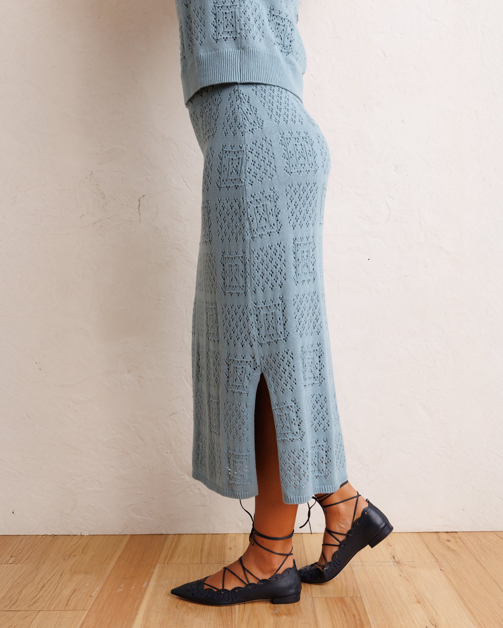 A person wearing a light blue open-knit midi skirt with a side slit, paired with black lace-up flats, standing against a textured off-white wall on a light wood floor.