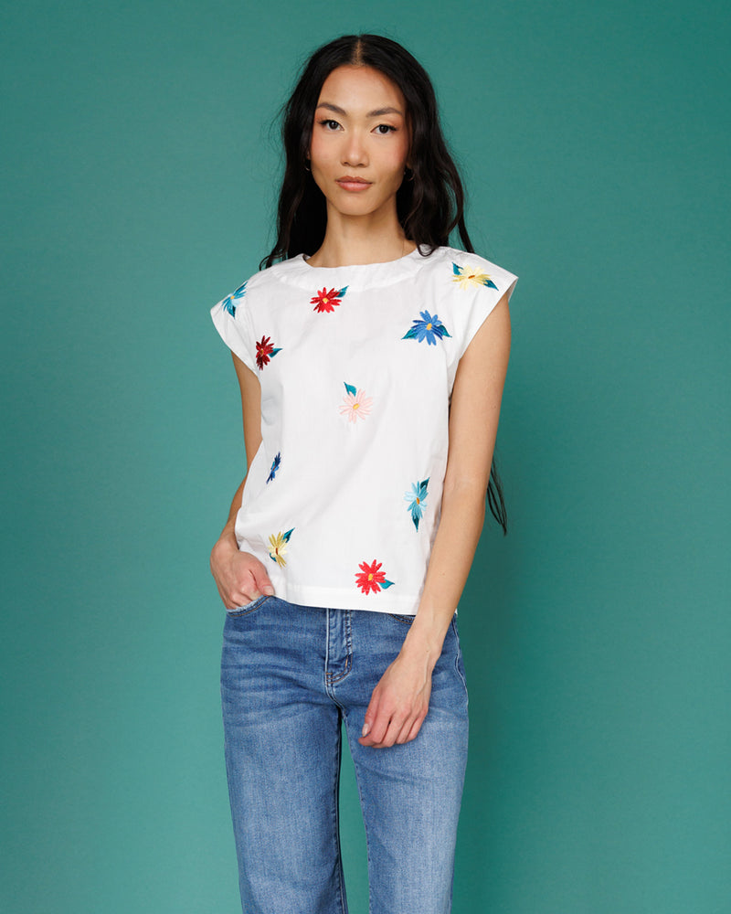 Woman wearing a white top with colorful floral patterns and blue jeans against a teal background