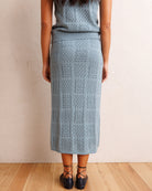 A woman shown from the back wearing a light blue-gray knit midi skirt with a textured geometric pattern, paired with a matching top and black lace-up shoes, standing against a beige wall on a wooden floor.