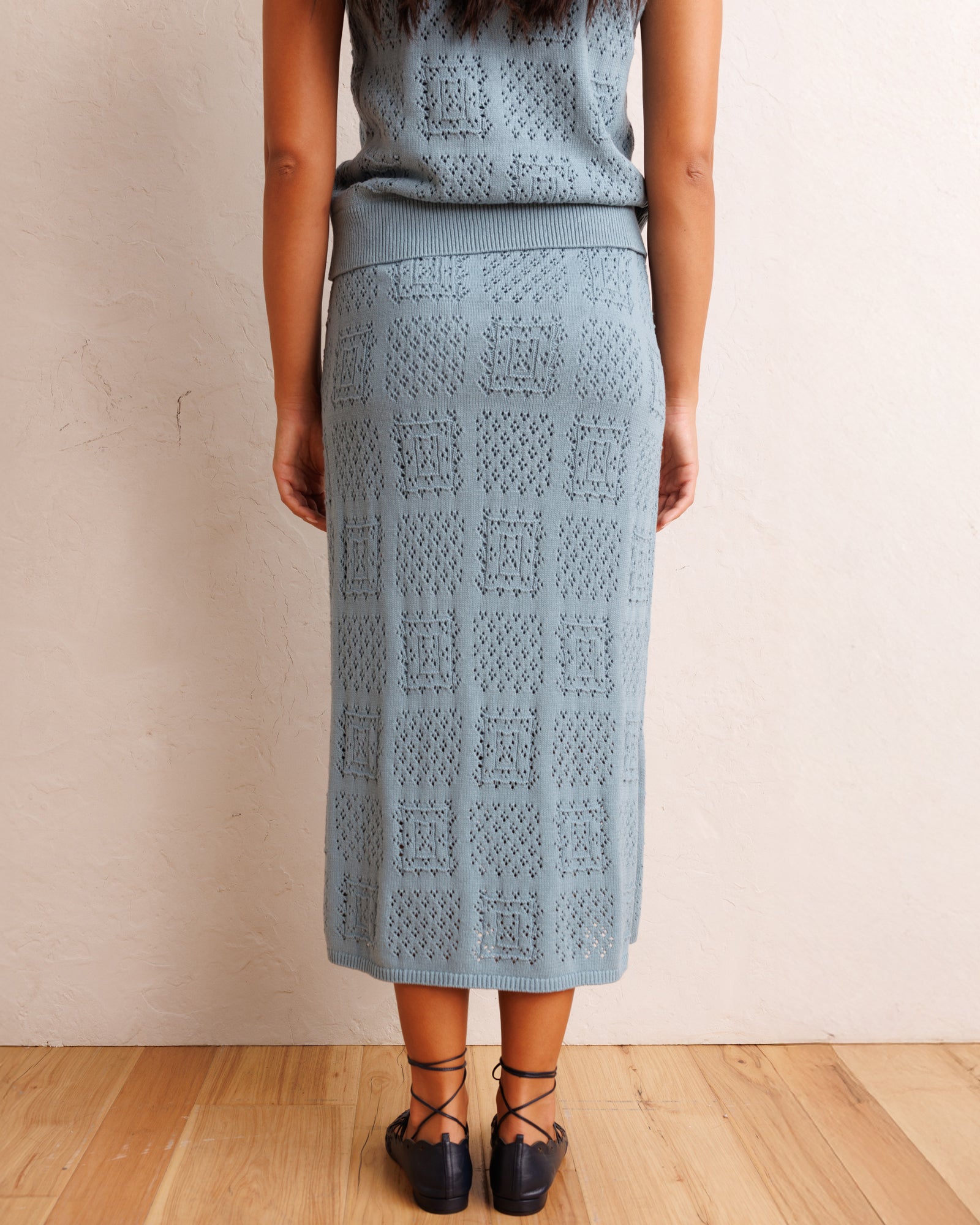A woman shown from the back wearing a light blue-gray knit midi skirt with a textured geometric pattern, paired with a matching top and black lace-up shoes, standing against a beige wall on a wooden floor.
