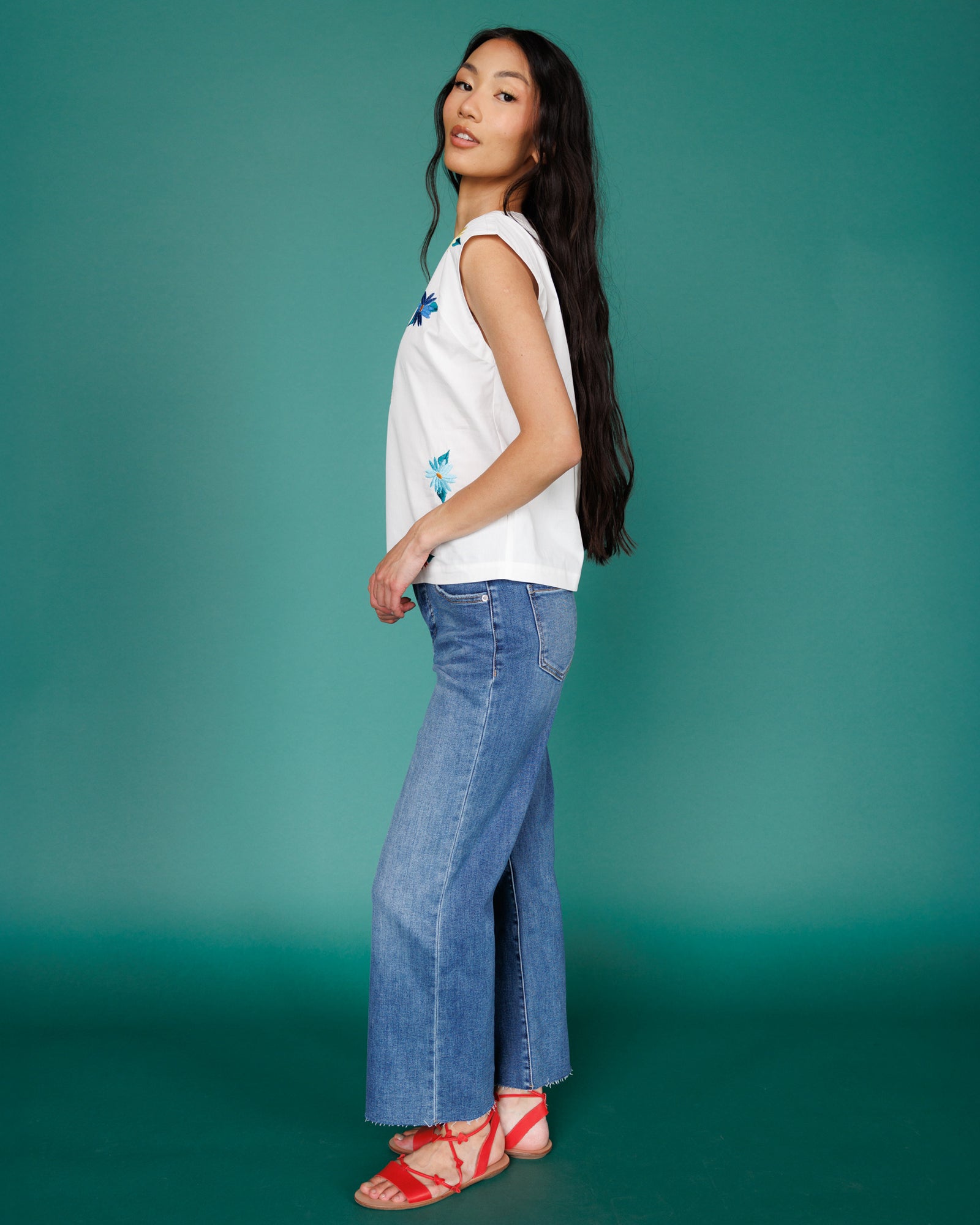 Woman wearing a white sleeveless top and blue jeans against a teal background