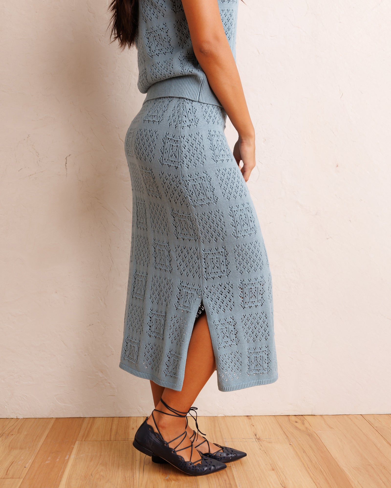 A woman wearing a light blue open-knit midi skirt with a side slit, paired with a matching top and dark lace-up flats, standing against a textured beige wall on a wood floor.