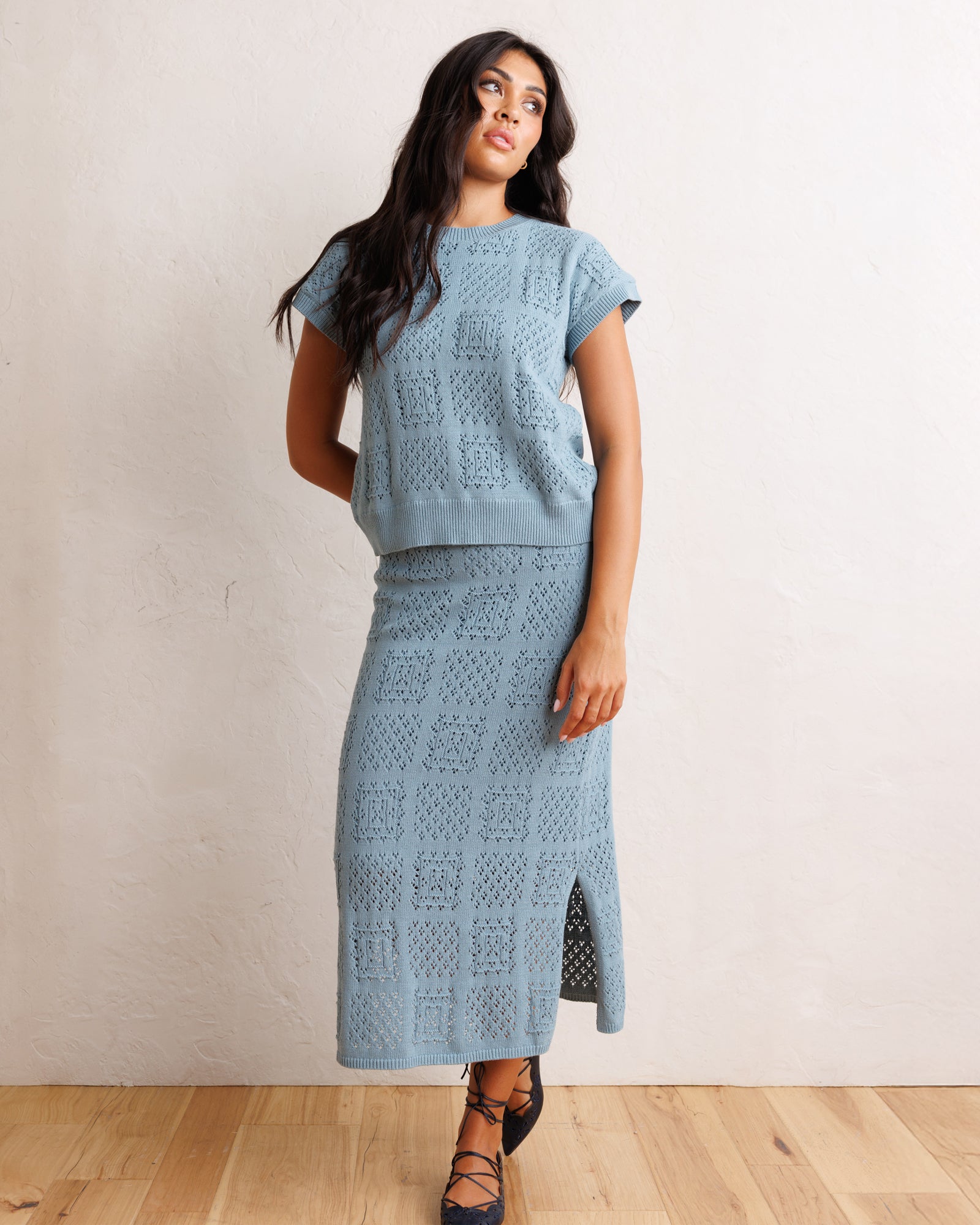 A woman modeling a light blue knit two-piece set consisting of a short-sleeve cropped top and a matching midi skirt with a side slit, set against a neutral off-white wall and light wood floor.