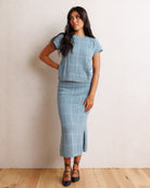 A woman models a light blue knit two-piece set consisting of a short-sleeve top and a matching midi skirt with a front slit, set against a neutral beige wall and light wood floor.