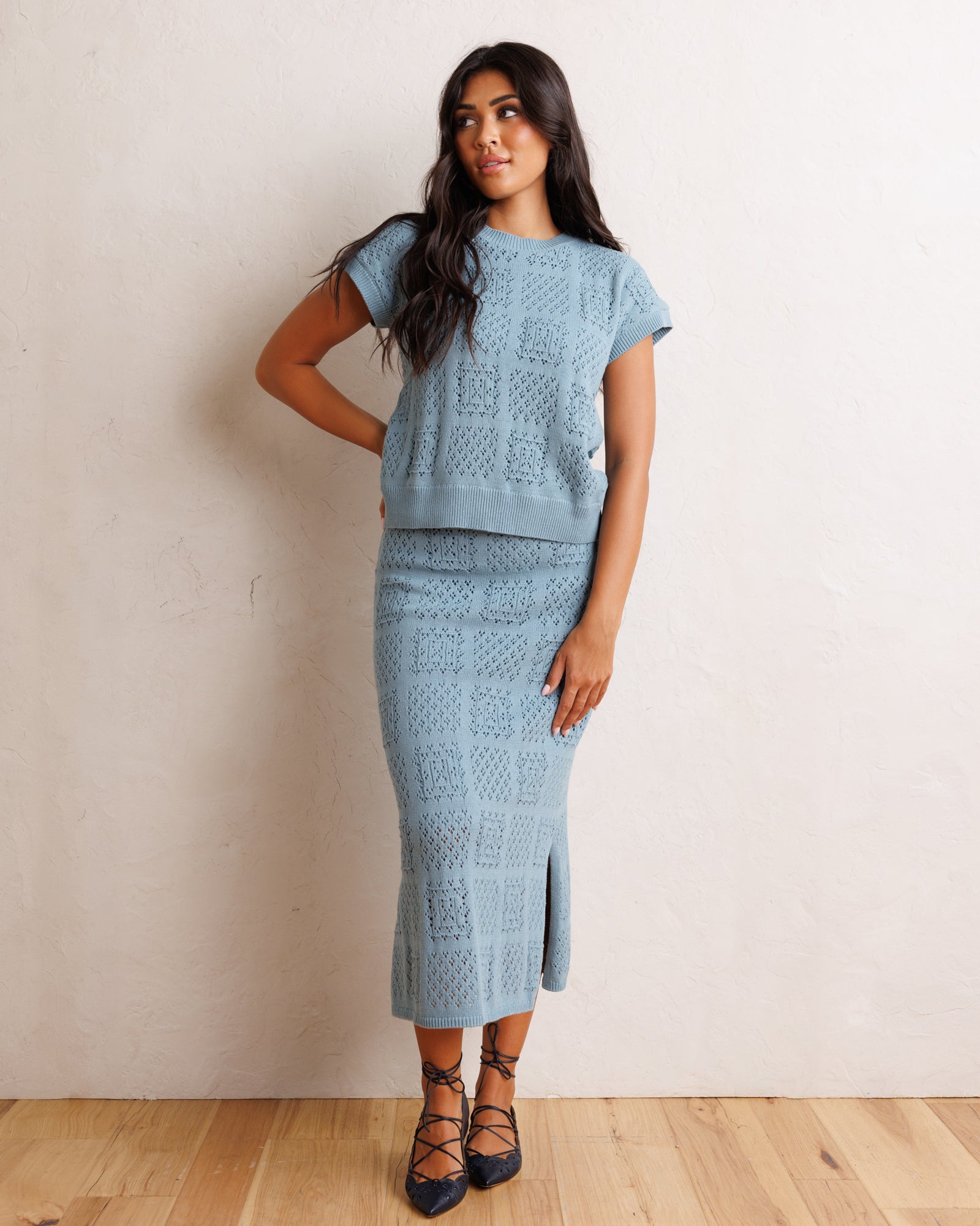 A woman models a light blue knit two-piece set consisting of a short-sleeve top and a matching midi skirt with a front slit, set against a neutral beige wall and light wood floor.