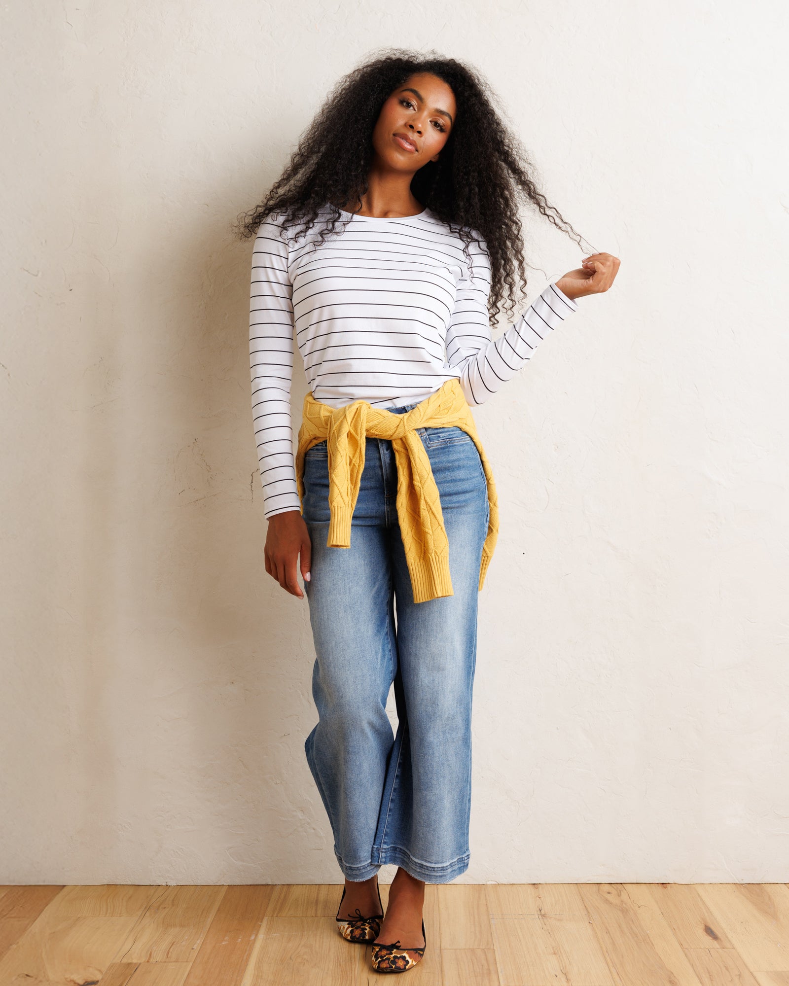 A woman stands against a textured off-white wall on a light wood floor, wearing a white long-sleeve top with thin navy horizontal stripes, light-wash blue jeans, and a bright yellow shirt tied around her waist. She is wearing leopard-print flat shoes.