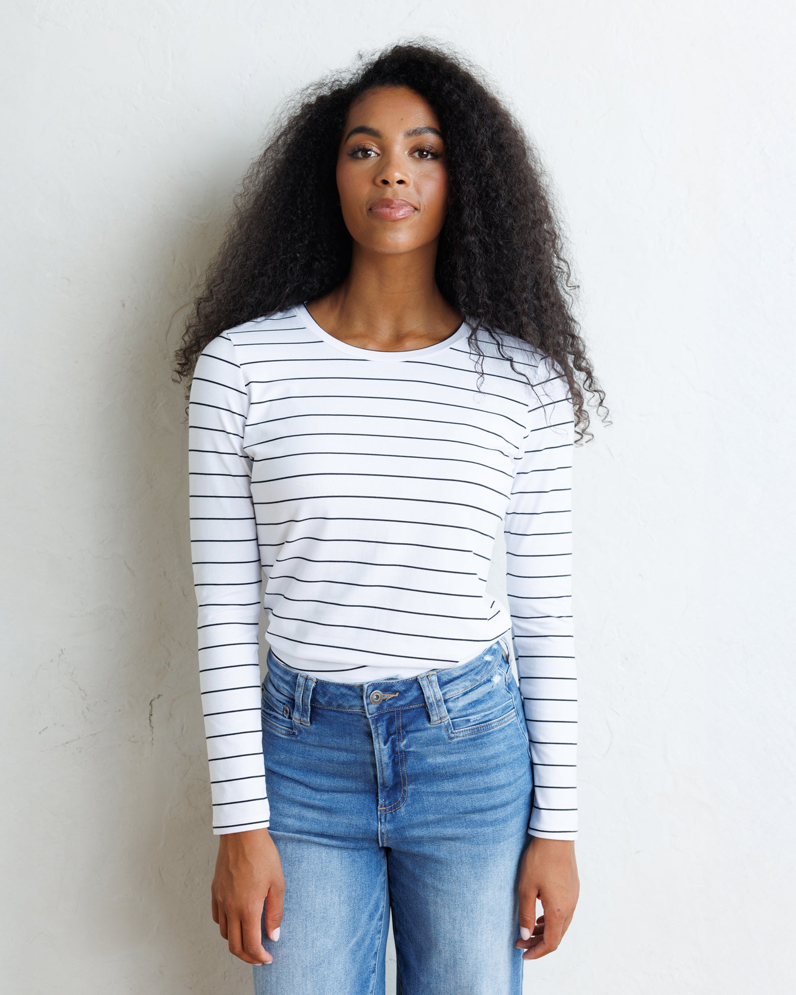 A woman with long, curly black hair stands against a light off-white wall, wearing a white long-sleeve crewneck top with thin black horizontal stripes and blue jeans.