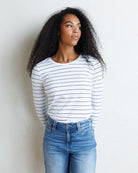 A woman with long curly black hair stands against a textured off-white wall, wearing a white long-sleeve crewneck top with thin black horizontal stripes and blue jeans