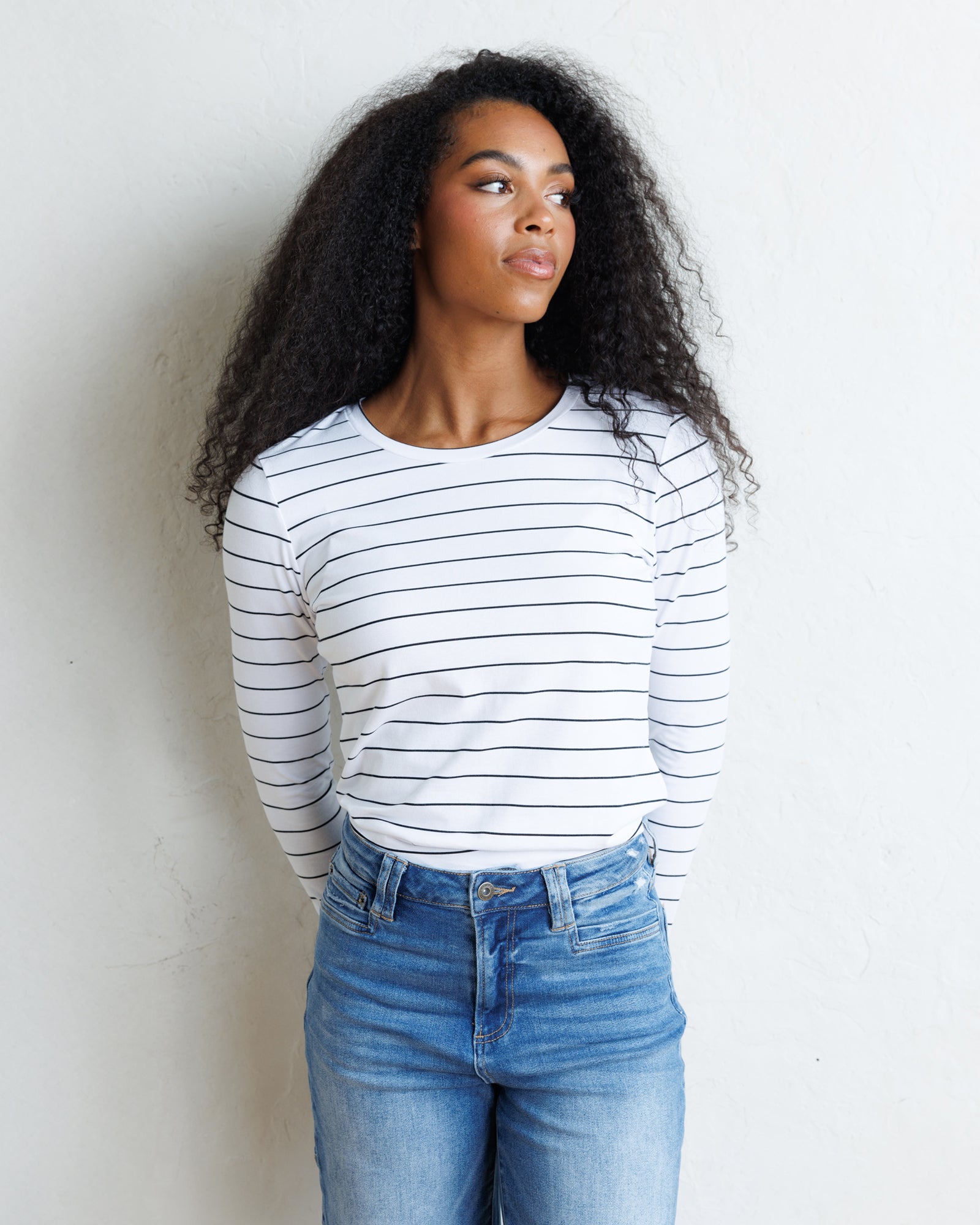 A woman with long curly black hair stands against a textured off-white wall, wearing a white long-sleeve crewneck top with thin black horizontal stripes and blue jeans
