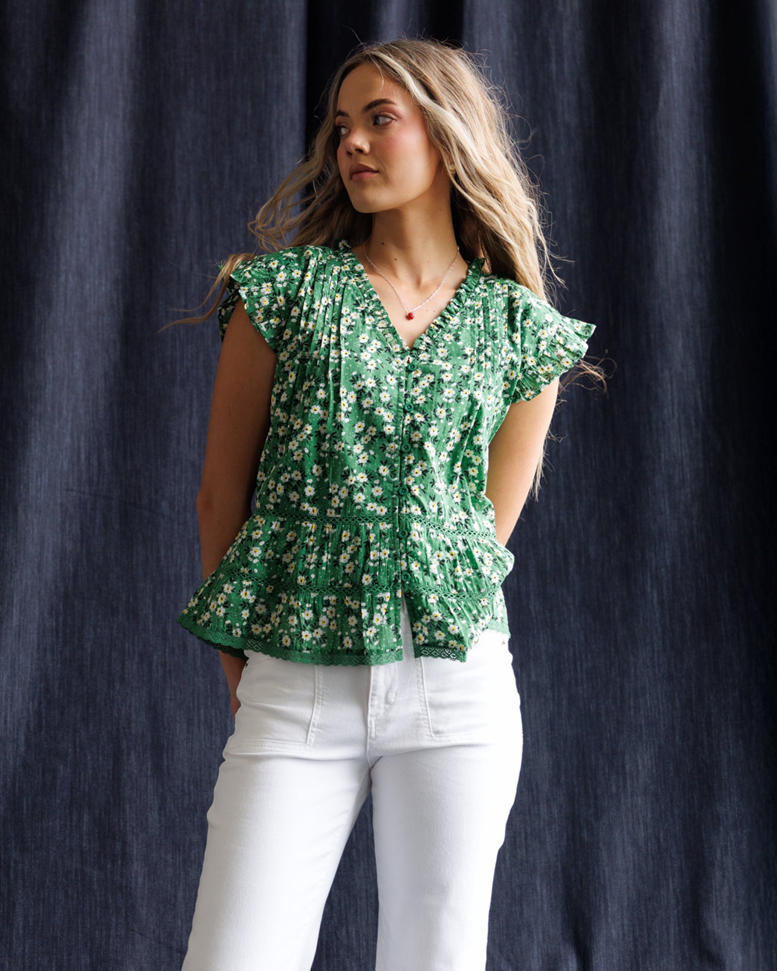 Woman wearing a green floral blouse and white pants against a dark curtain background