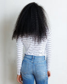A person shown from behind wearing a white long-sleeve shirt with thin black horizontal stripes and light blue jeans, standing against a plain off-white wall.