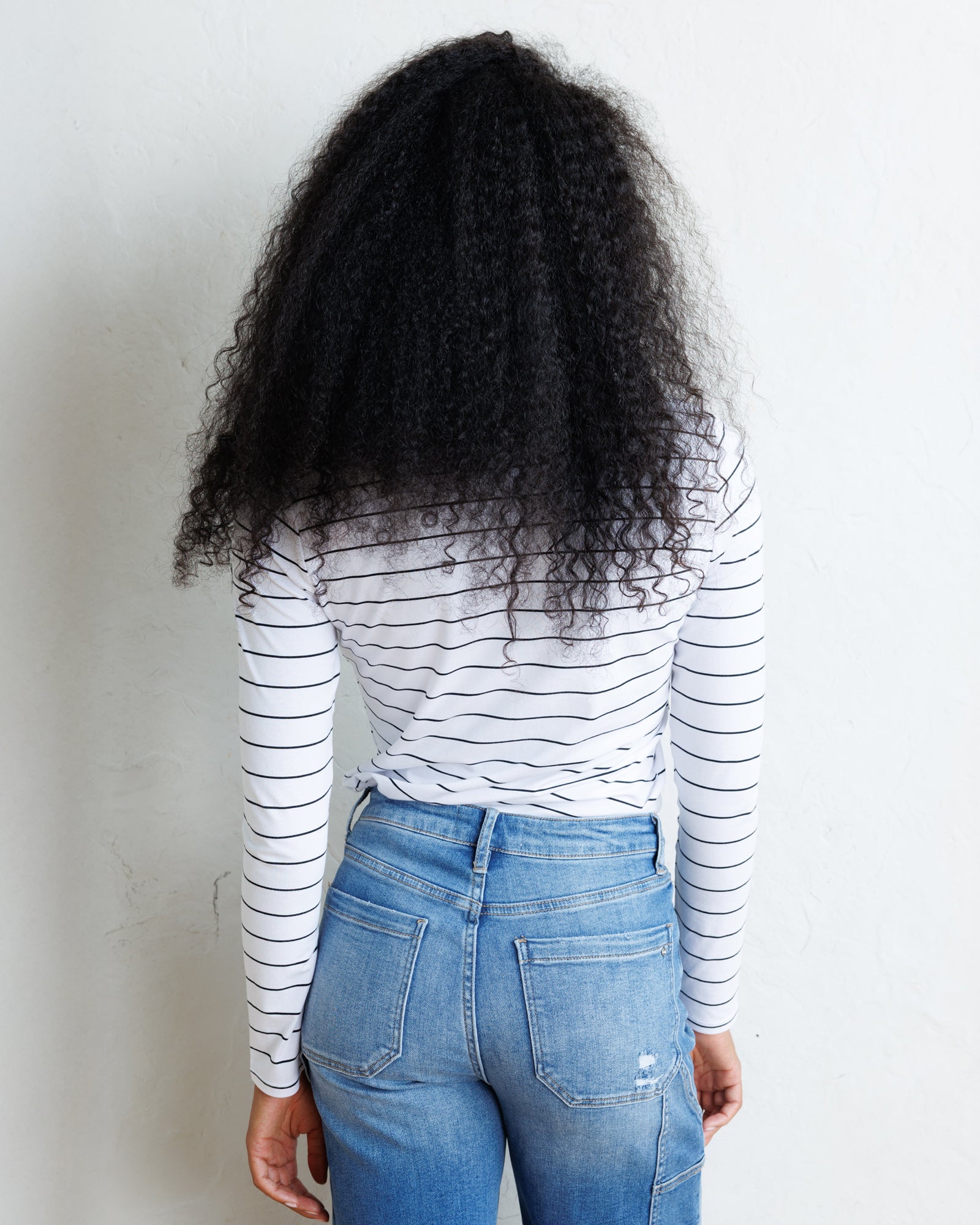 A person shown from behind wearing a white long-sleeve shirt with thin black horizontal stripes and light blue jeans, standing against a plain off-white wall.