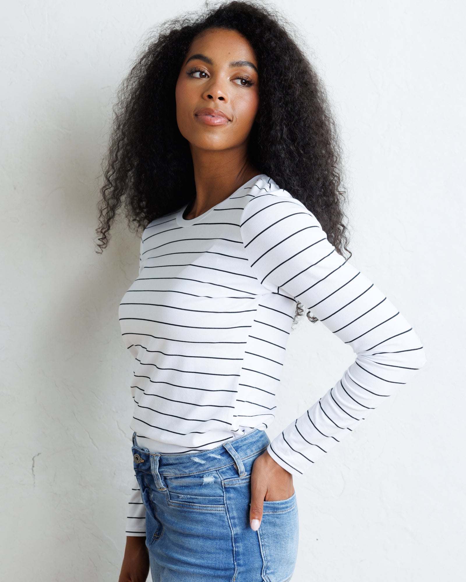 A woman with curly black hair wearing a white long-sleeve crewneck top with thin black horizontal stripes and blue jeans, standing against a white wall.