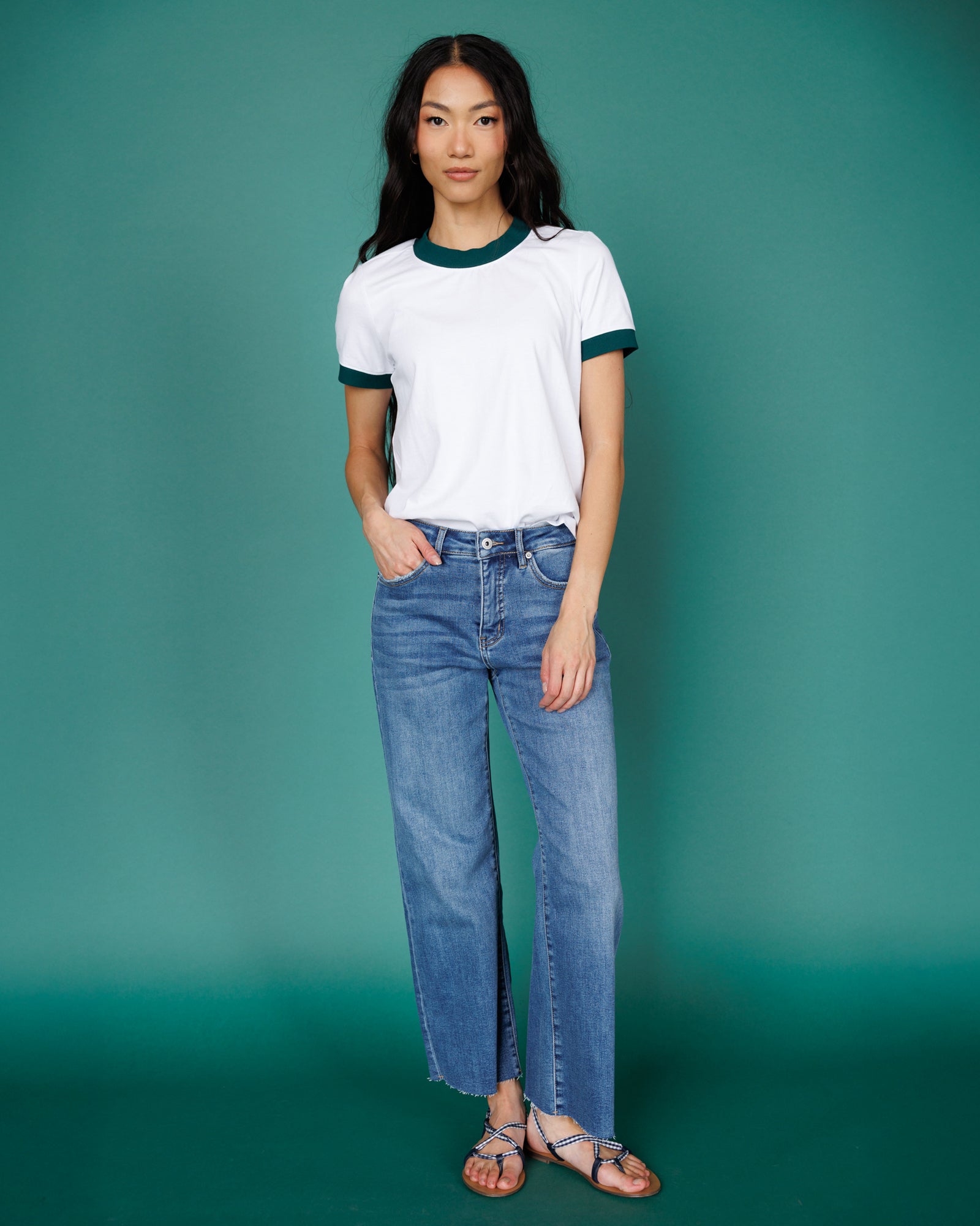 Woman wearing a white t-shirt with green accents and blue jeans against a teal background