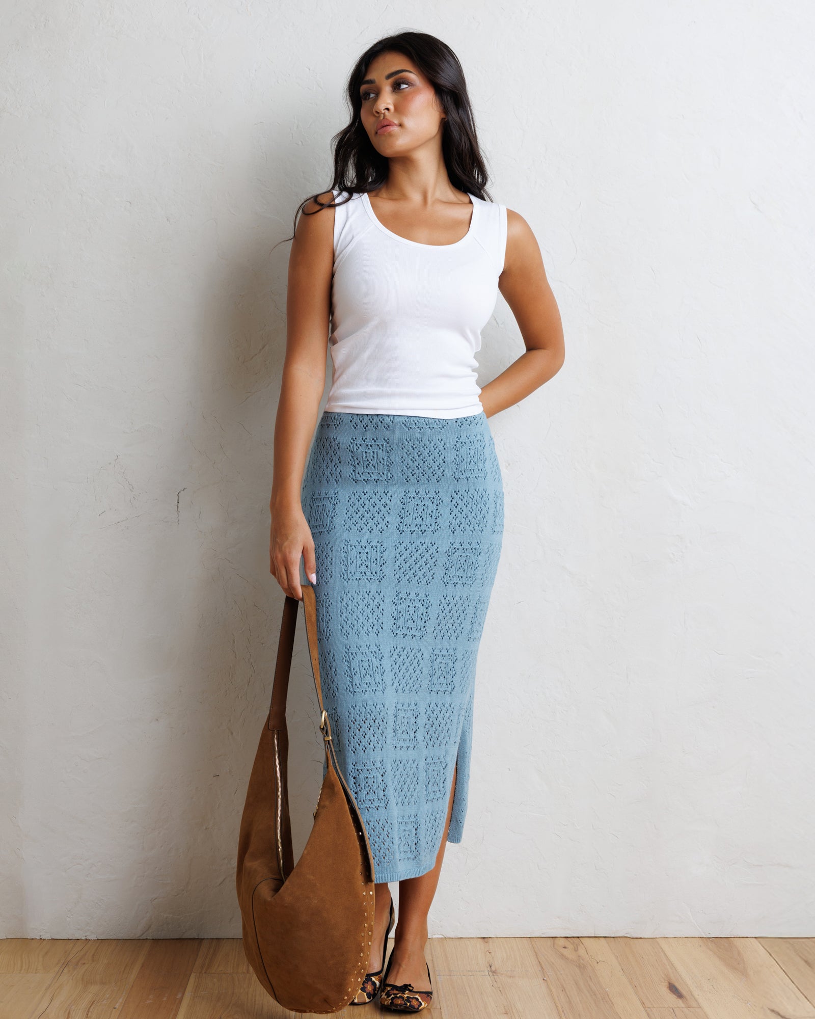 A woman stands against a textured off-white wall wearing a white tank top and a light blue crocheted midi skirt with a side slit. She is holding a large brown suede shoulder bag.