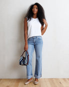 A woman stands against a textured off-white wall on a light wood floor, wearing a white sleeveless top, light-wash blue jeans with rolled cuffs, and beige flat shoes. She holds a small striped handbag.