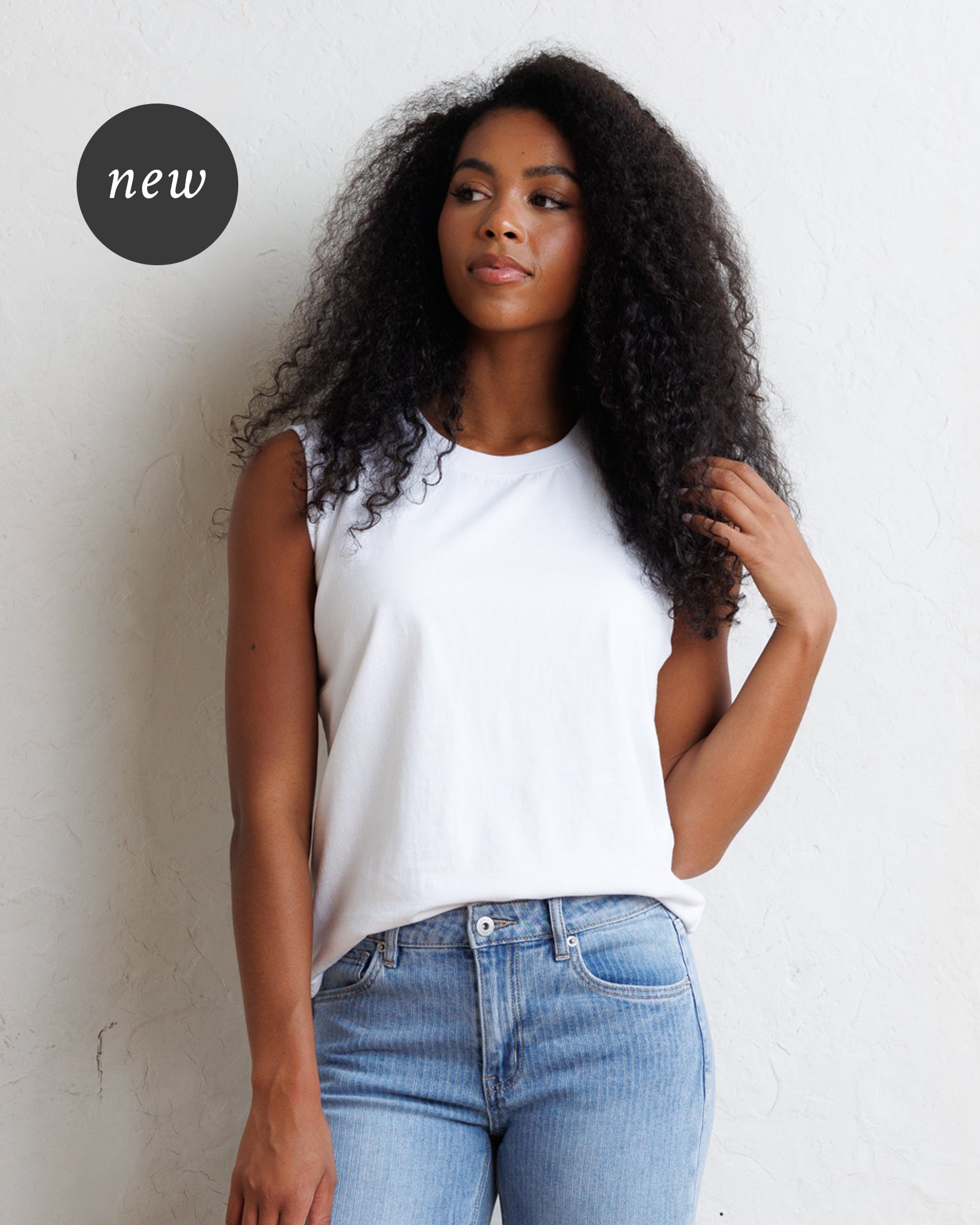 A woman with long, curly black hair wearing a white sleeveless top and light blue jeans, standing against a textured off-white wall.