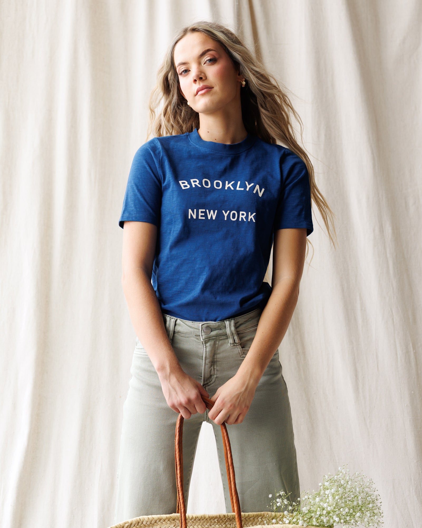 Woman wearing a blue 'Brooklyn New York' t-shirt against a white curtain background