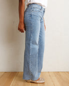 A person wearing light-wash blue wide-leg jeans, a white top, and beige woven flat shoes, standing against a textured off-white wall on a light wood floor.
