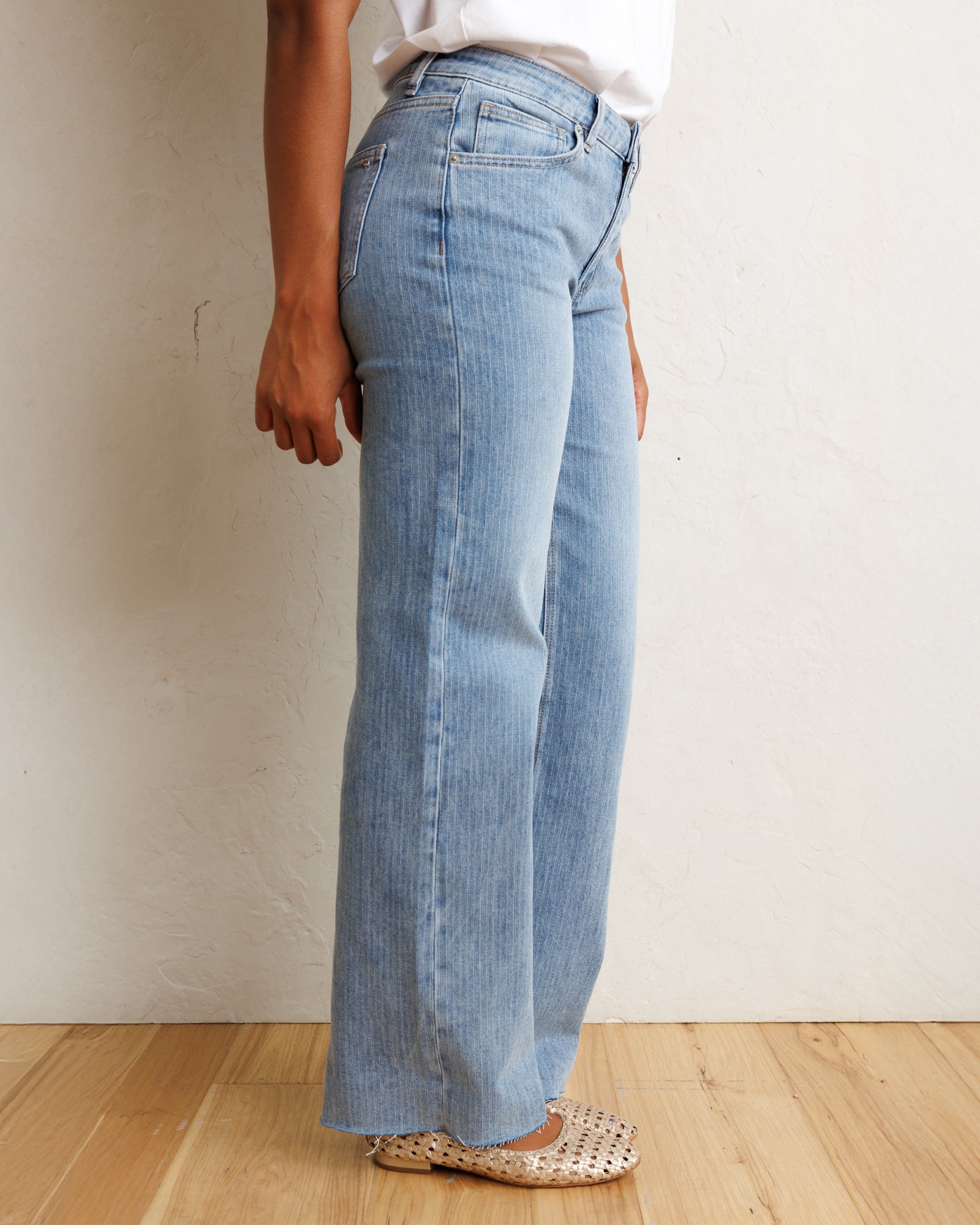 A person wearing light-wash blue wide-leg jeans, a white top, and beige woven flat shoes, standing against a textured off-white wall on a light wood floor.