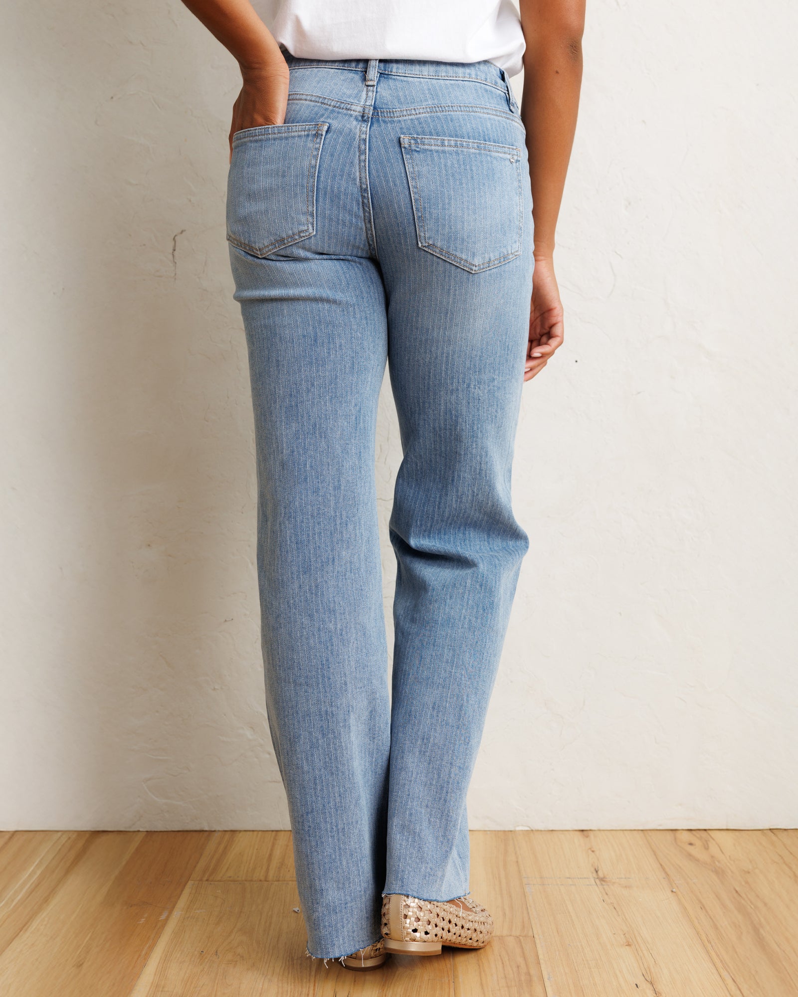 A person shown from the back wearing light-wash blue jeans, a white top, and beige flat shoes, standing against a textured off-white wall on a light wood floor.
