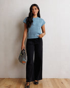 A woman stands against a textured off-white wall on a light wood floor, wearing a light blue short-sleeve knit top with a geometric pattern, black wide-leg pants, and black polka-dot flat shoes, while holding a gray patterned handbag.