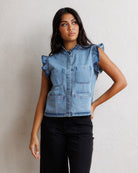 A woman modeling a light blue denim sleeveless vest with ruffled cap sleeves, a button front, and front patch pockets, paired with black pants against a textured off-white wall.