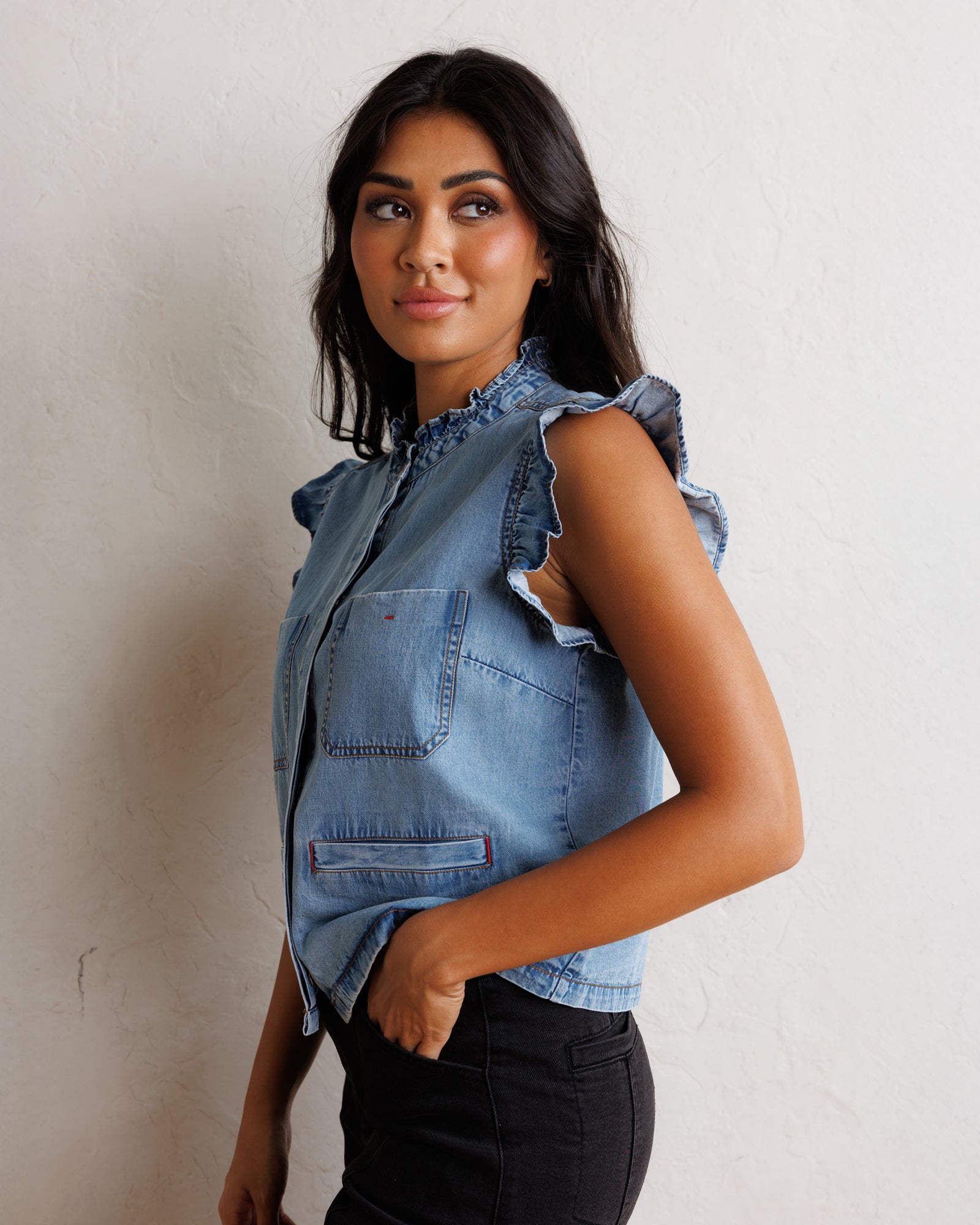 A woman wearing a sleeveless light blue denim vest with ruffled trim and front pockets, paired with black pants, standing against a textured off-white wall.