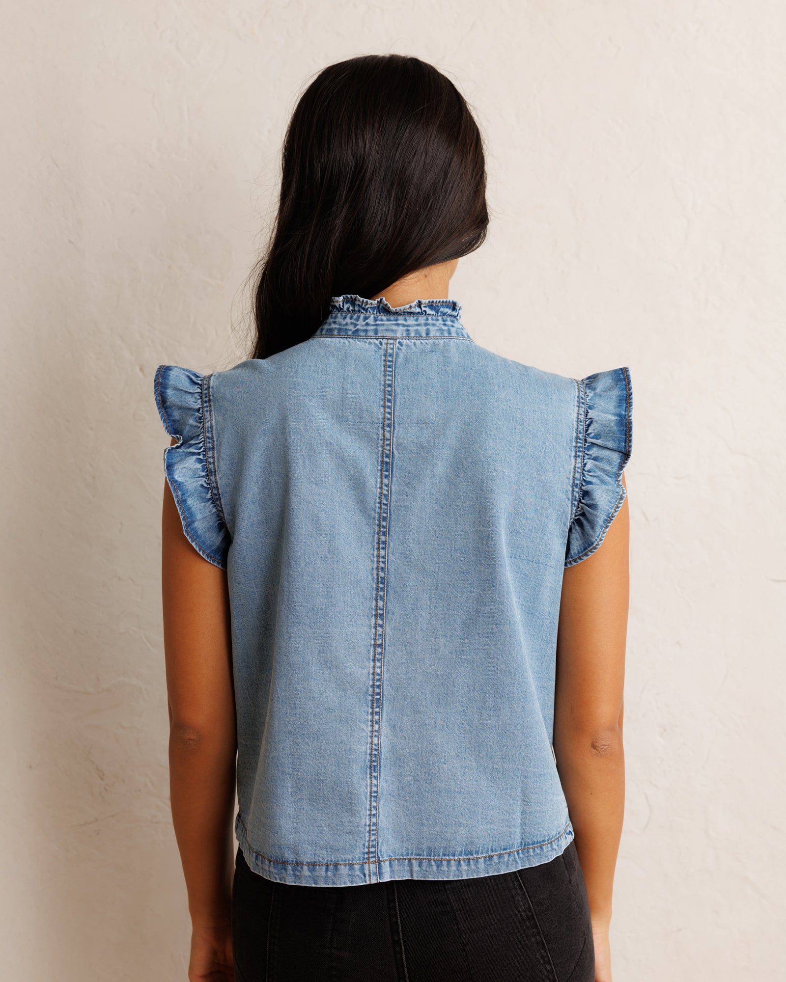 A woman viewed from the back wearing a light blue denim sleeveless top with ruffled cap sleeves and a high ruffled collar, paired with black pants against a textured off-white wall.