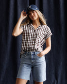 Woman wearing a checkered shirt, denim shorts, and a cap against a dark background