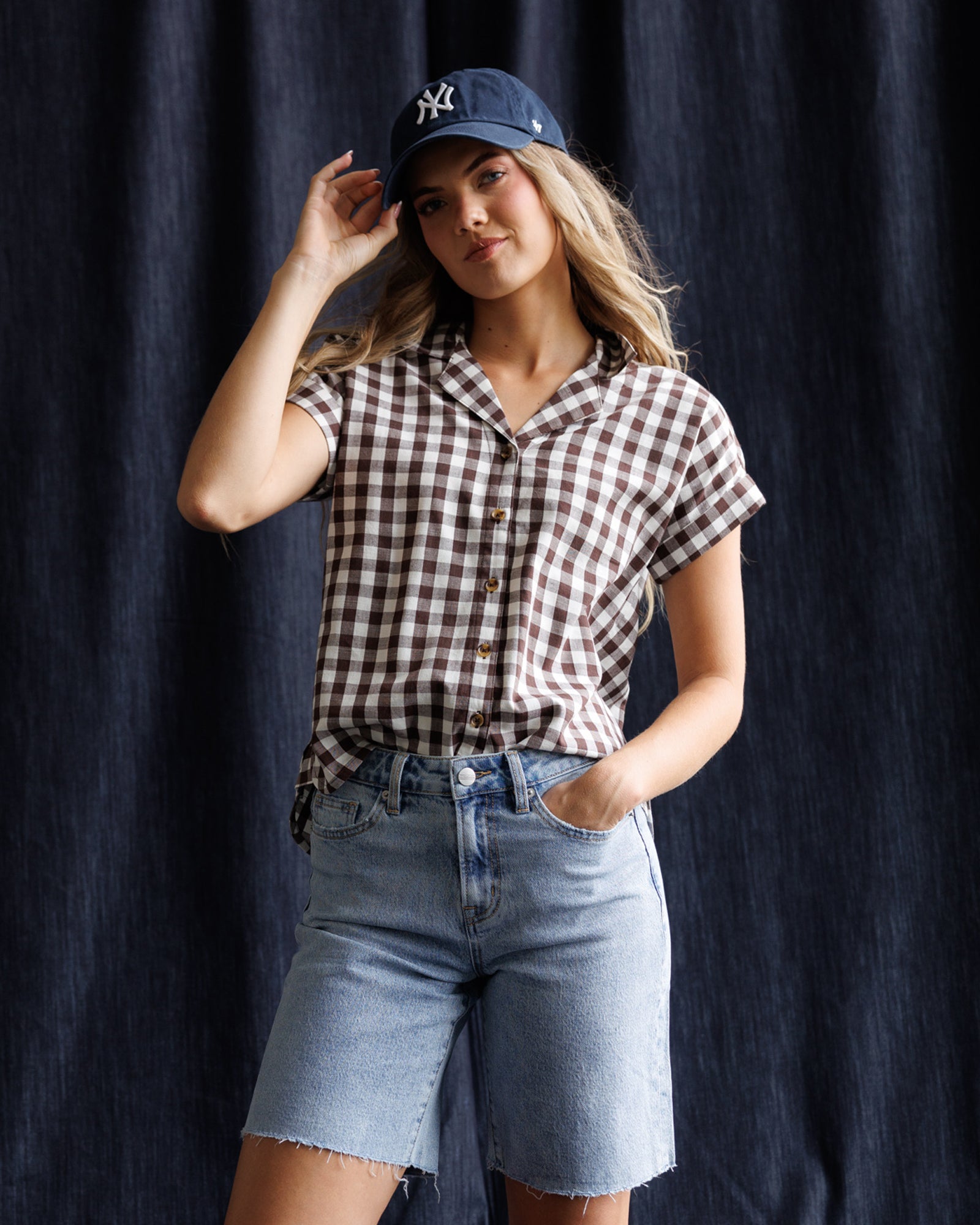 Woman wearing a checkered shirt, denim shorts, and a cap against a dark background