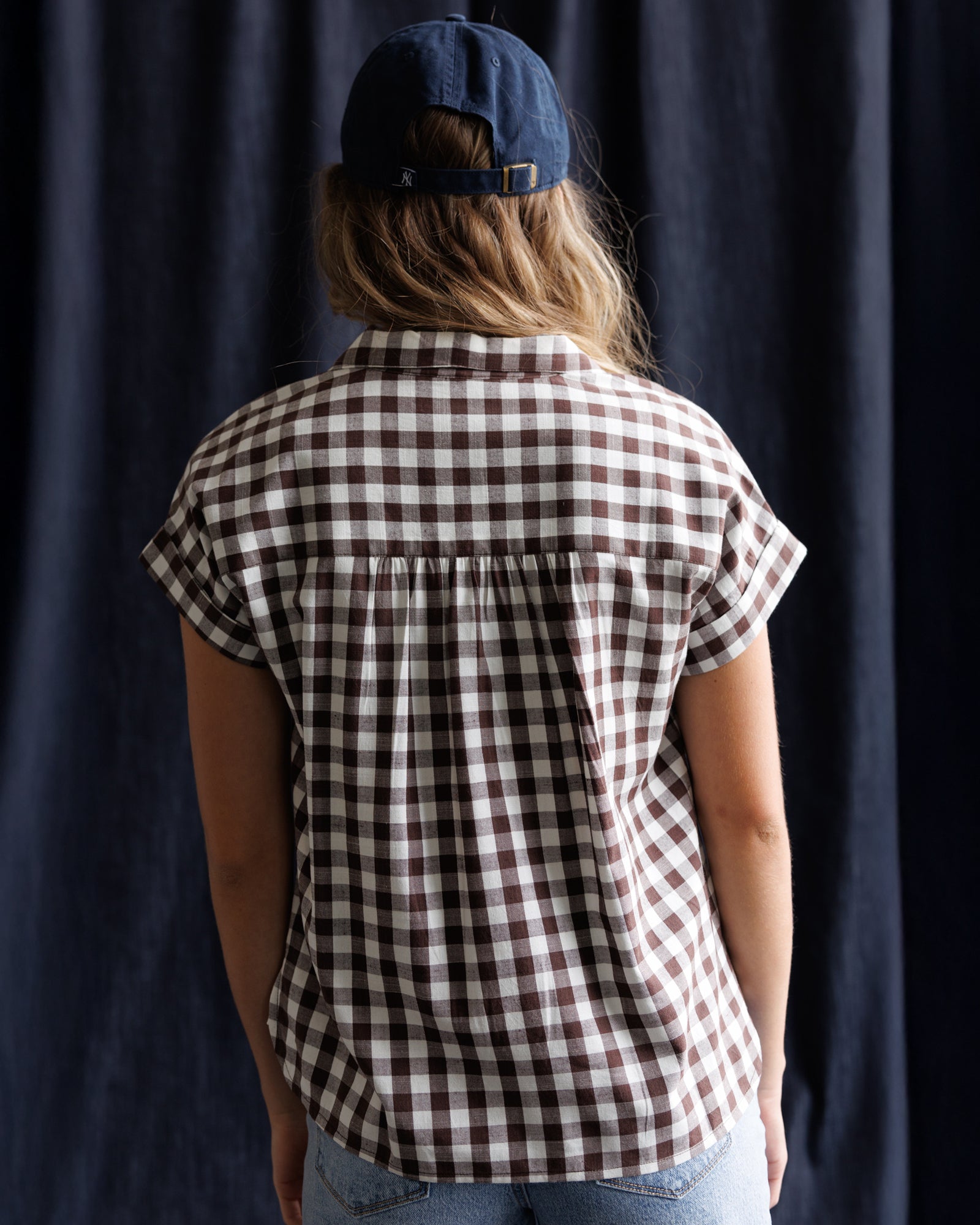 Person wearing a brown and white checkered shirt with a blue cap against a dark curtain background