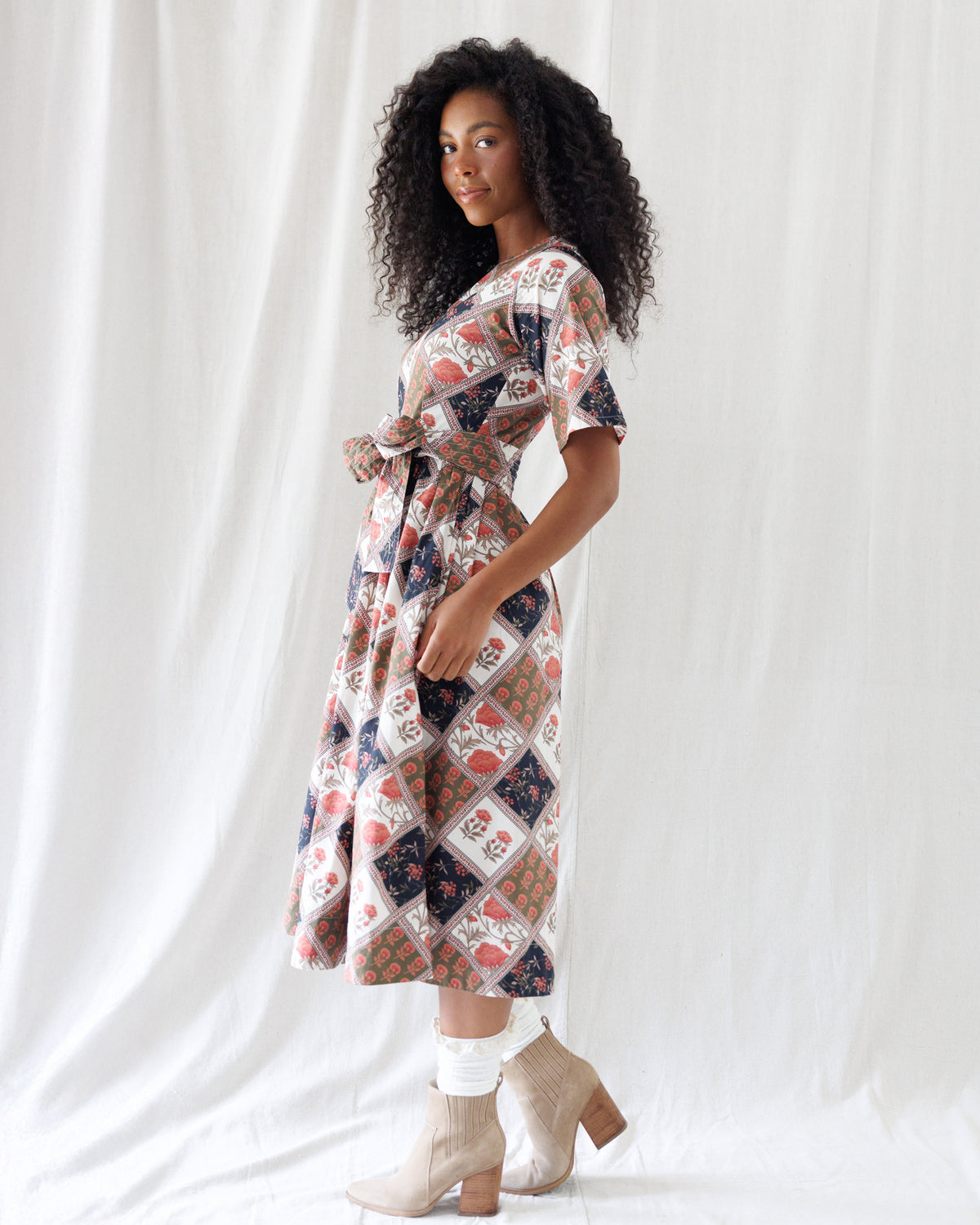 Side profile showing the relaxed elbow-length sleeves, defined waist with front bow tie, and breezy calf-length midi hemline of the patterned patchwork dress.