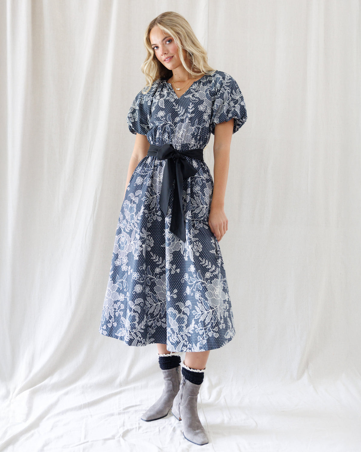 A woman modeling a black midi dress with a white botanical pattern, featuring puff sleeves, a V-neckline, and a matching navy tie belt at the waist. She is wearing gray suede ankle boots with black socks, standing against a draped white fabric backdrop.