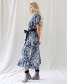 A woman modeling a short-sleeve midi dress with a black and white floral pattern and a black waist tie, standing against a white draped fabric backdrop.
