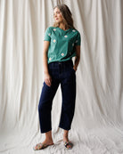Woman wearing a green floral t-shirt and dark blue jeans against a white curtain background