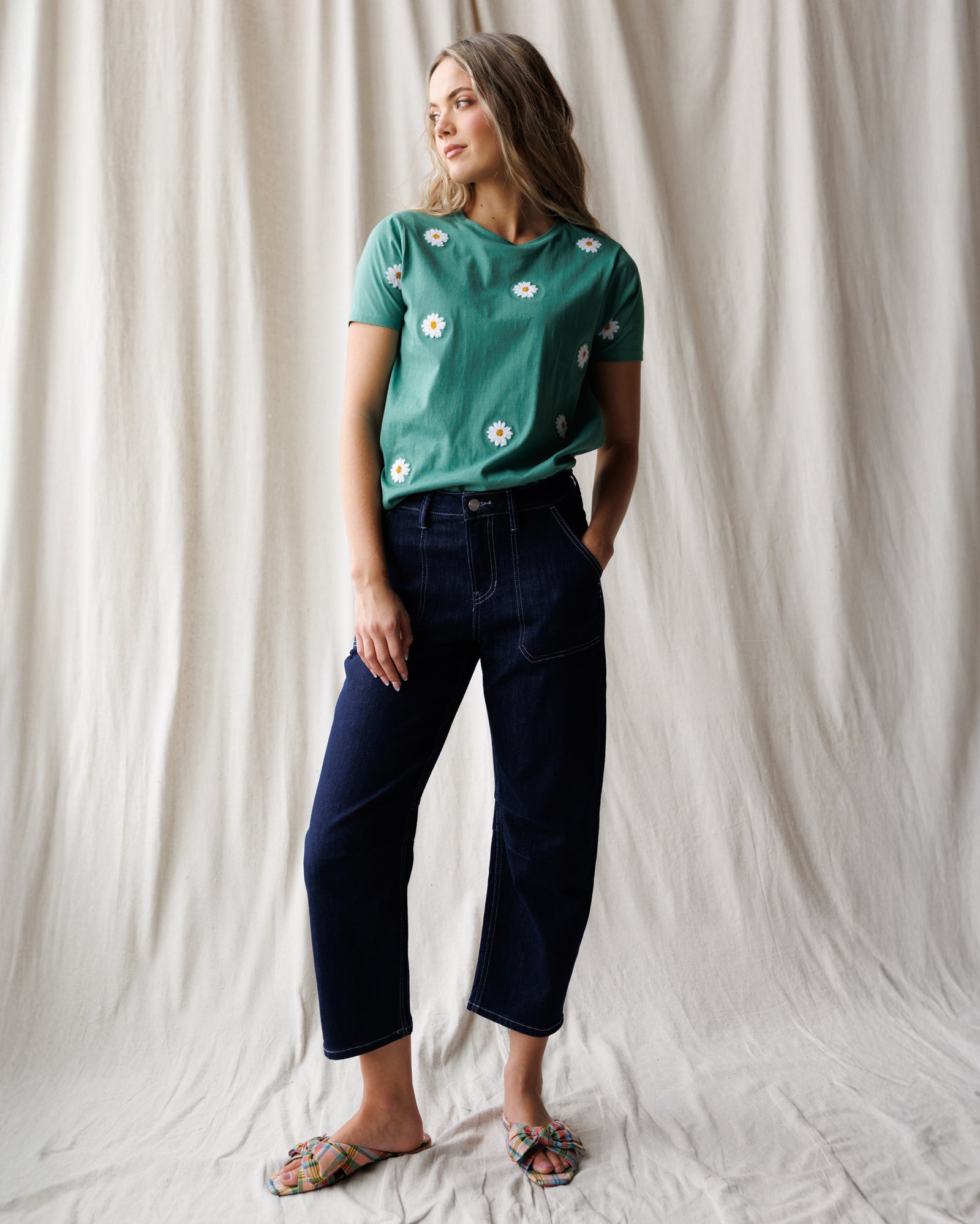 Woman wearing a green floral t-shirt and dark blue jeans against a white curtain background