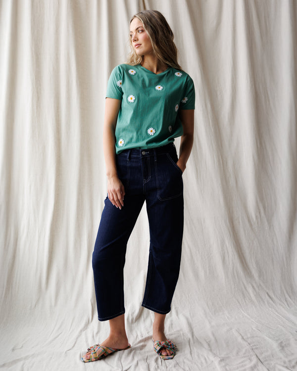 Woman wearing a green floral t-shirt and dark blue jeans against a white curtain background