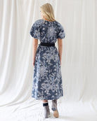 Back view of a woman modeling a short-sleeve midi dress with a black and white floral pattern and a black waist tie, standing against a white draped fabric backdrop.