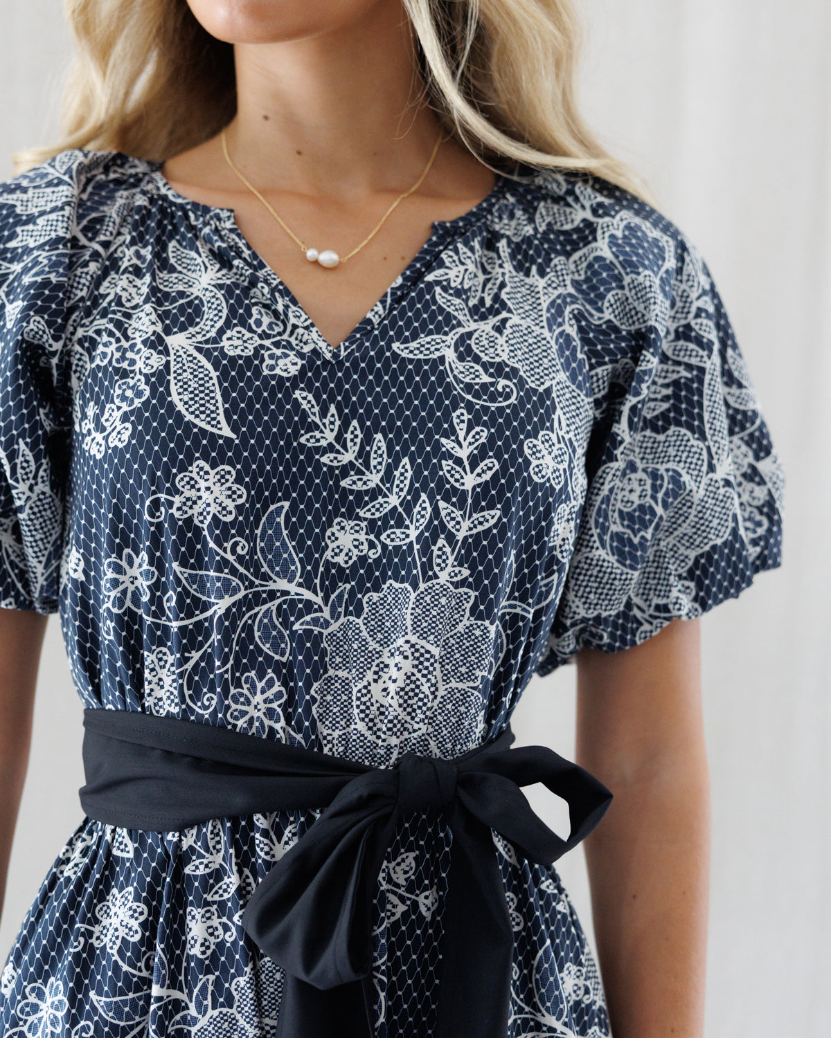 A close-up of a woman wearing a black blue and white floral print dress with a matching fabric tie belt at the waist. The dress features a V-neckline, short puff sleeves, and a delicate white polka-dot pattern throughout.