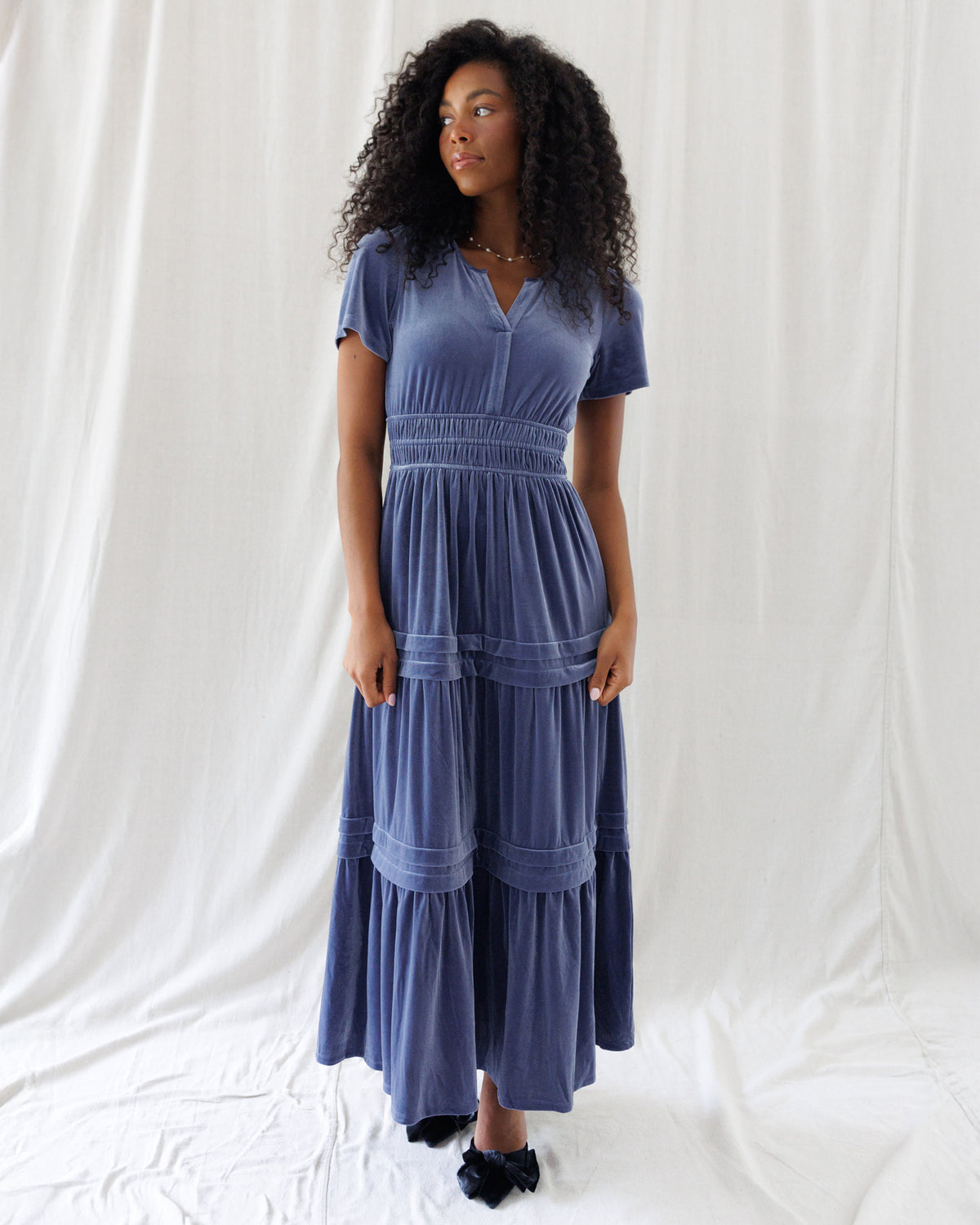 Front view of a dusty slate blue velvet maxi dress featuring short sleeves, a split V-neckline, wide smocked elastic waist, and tiered skirt with horizontal tuck detailing.